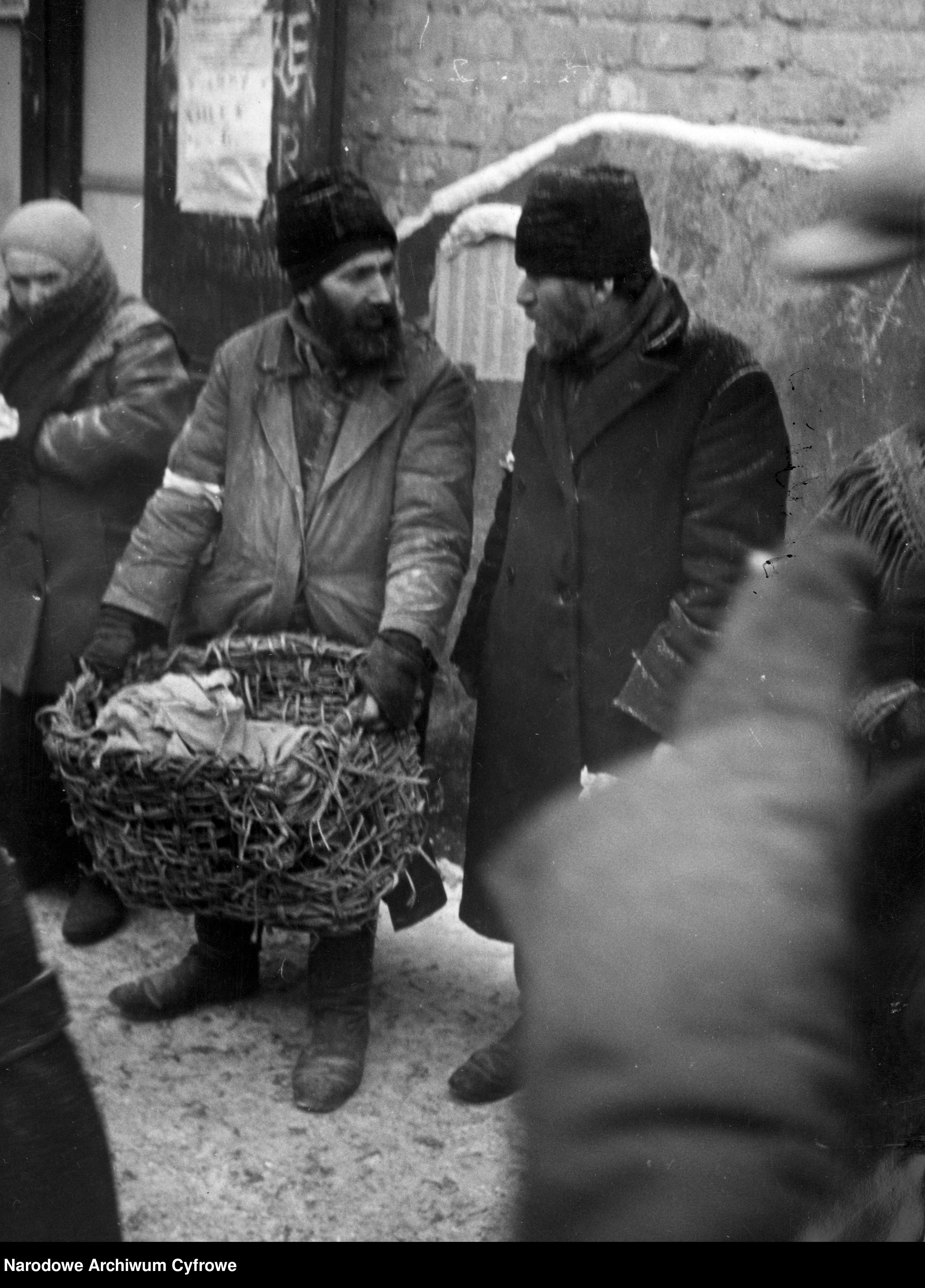 Żydzi w warszawskim getcie. Dwóch mężczyzn, jeden trzyma kosz z jasną tkaniną w środku. 