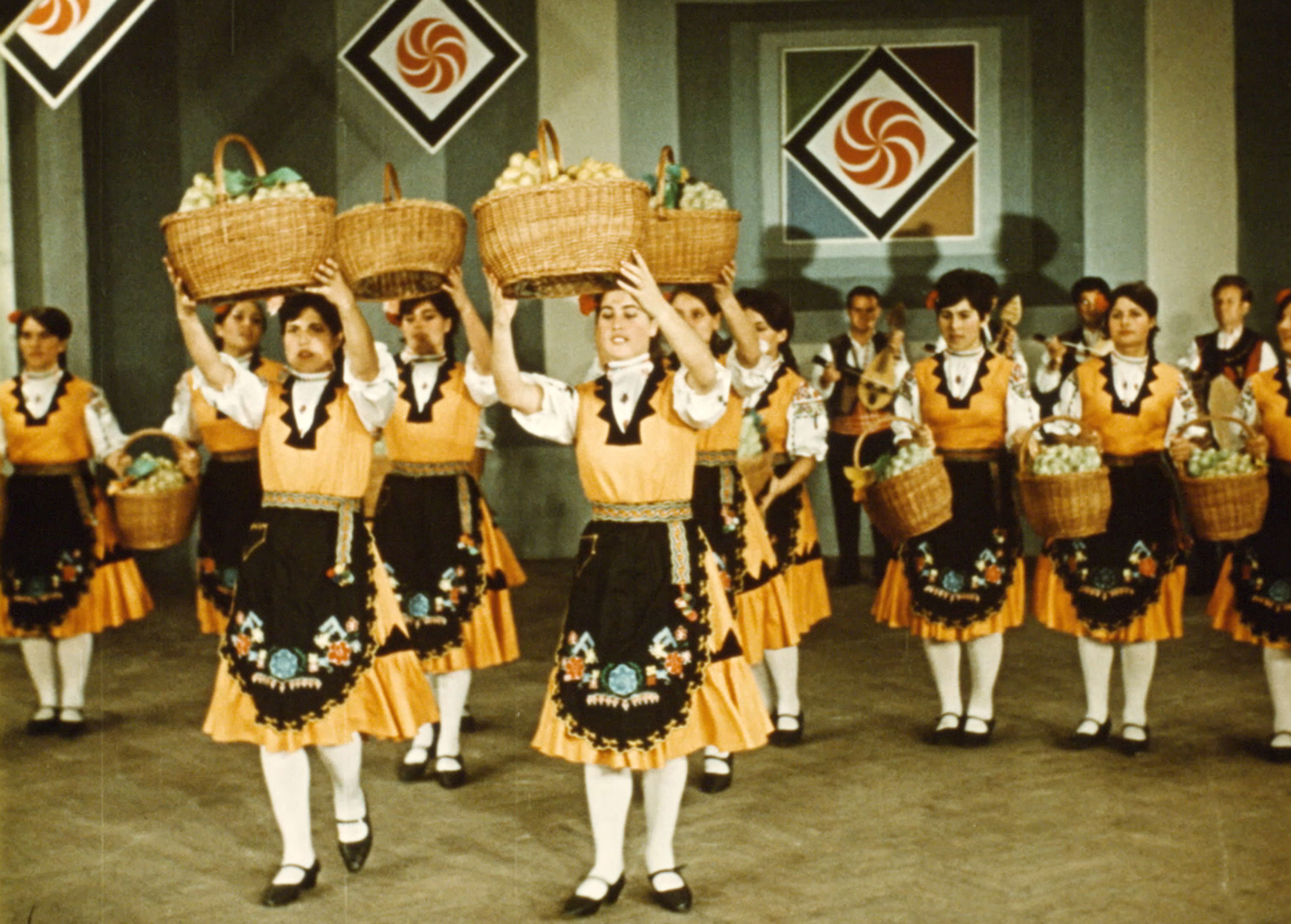 Grupa tancerek z trzyma koszyki pełne owoców nad głową. 
