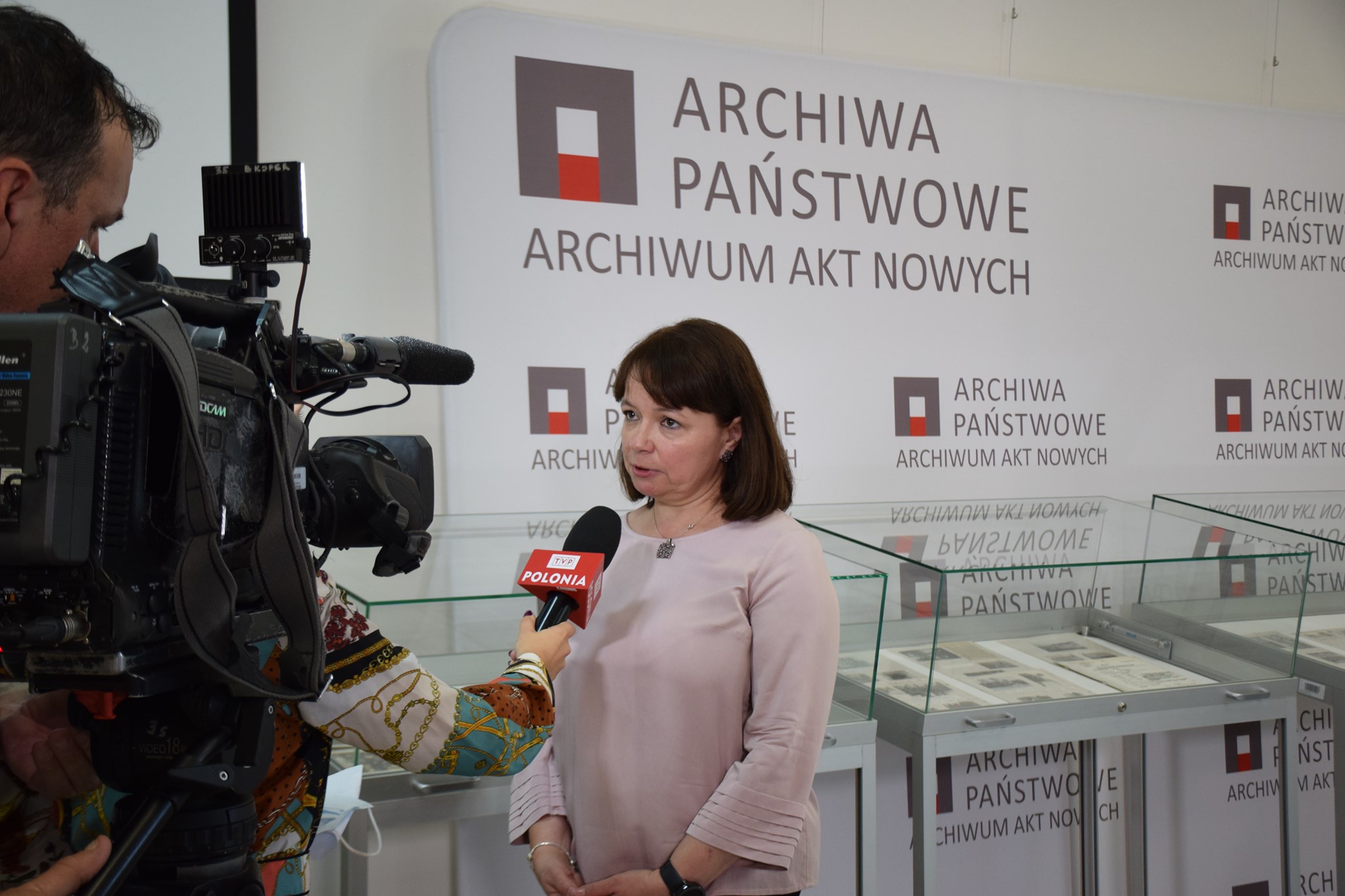 Dyrektor Lucyna Harc podczas wywiadu dla TVP Polonia. W tle szklane gabloty z materiałami archiwalnymi.