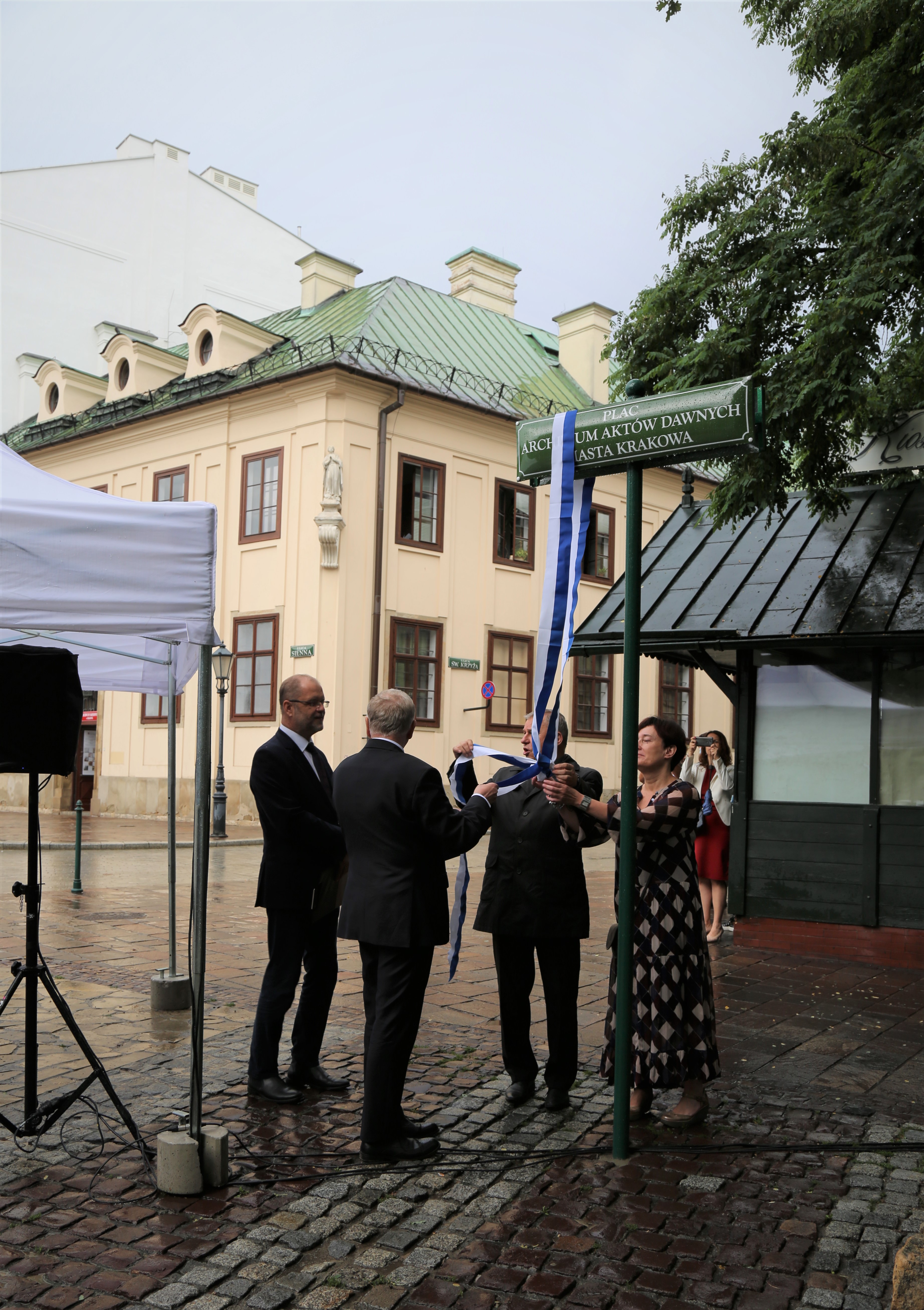 Cztery osoby trzymają biało-niebieską szarfę zawieszoną na tabliczce z nazwą Plac Archiwum Aktów Dawnych Miasta Krakowa.