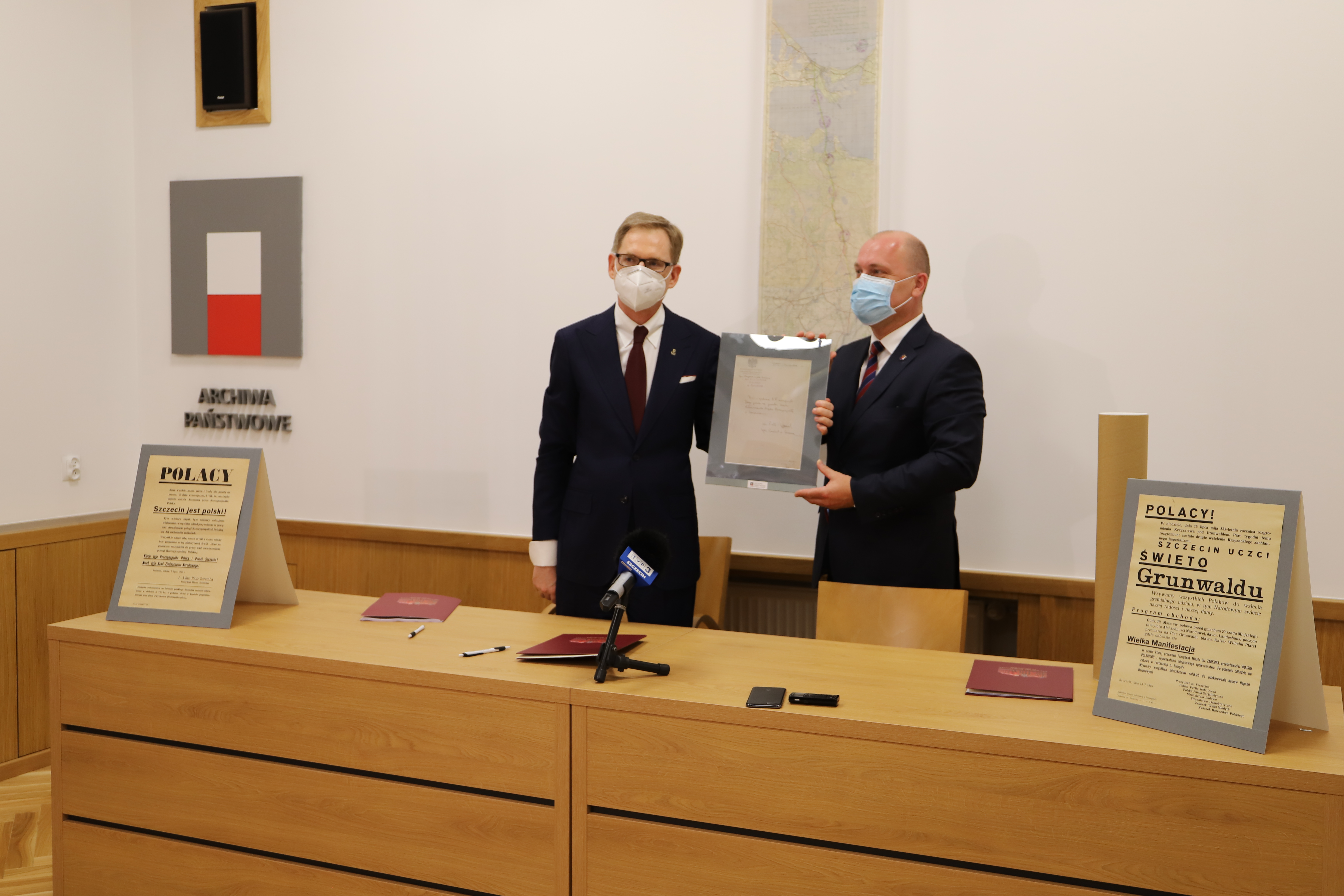 Prezydent Krystek i dyrektor Kowalczyk pozują z ramą z materiałem archiwalnym, w tle wisi mapa.