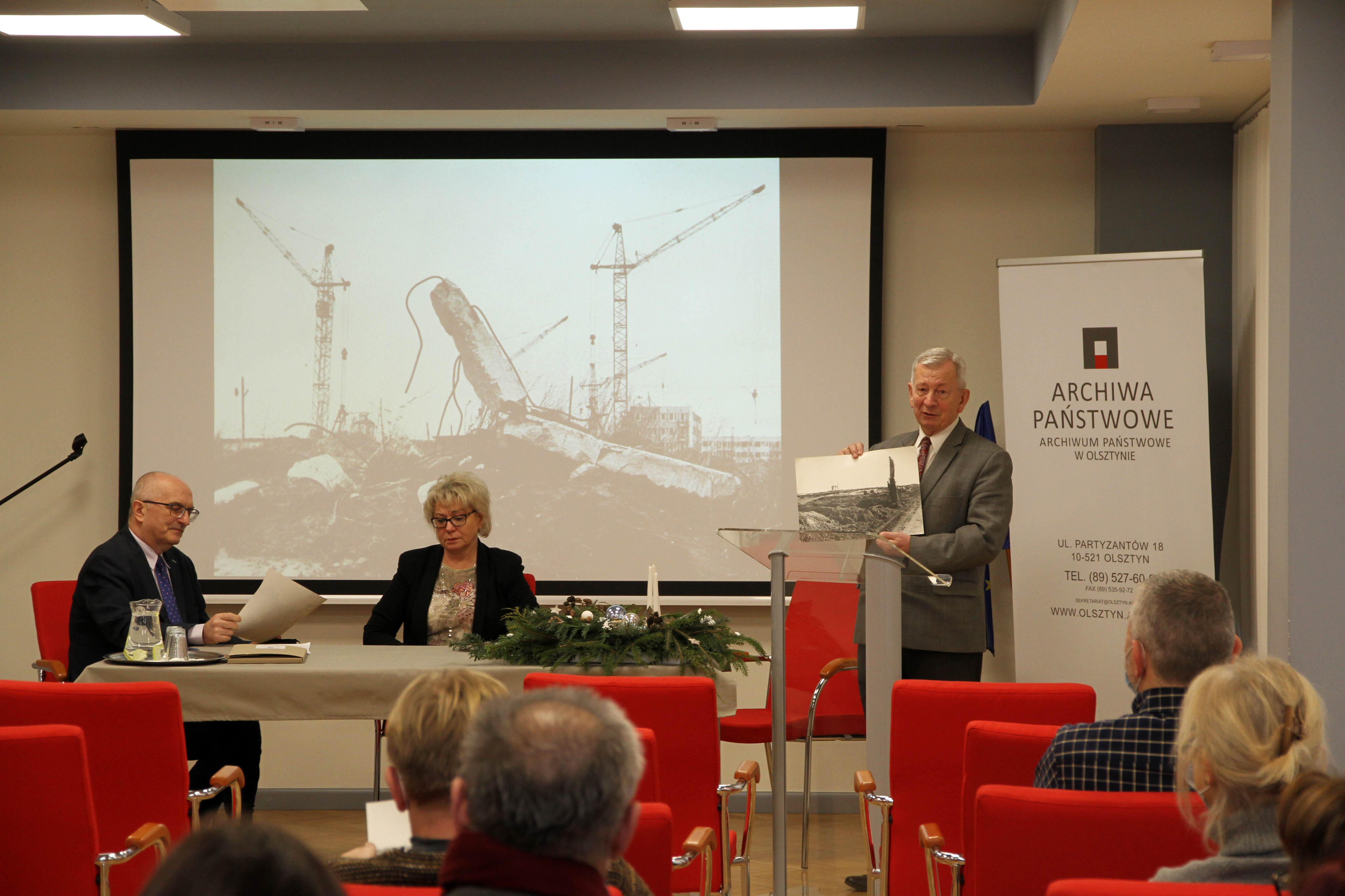 Dyrektor Kasparek i redaktor Gieczewska siedzą za stołem konferencyjnym. Po lewej stoi Roman Przedwojski i prezentuje jedno z przekazanych zdjęć. W tle prezentacja z innymi przekazanymi zdjęciami.