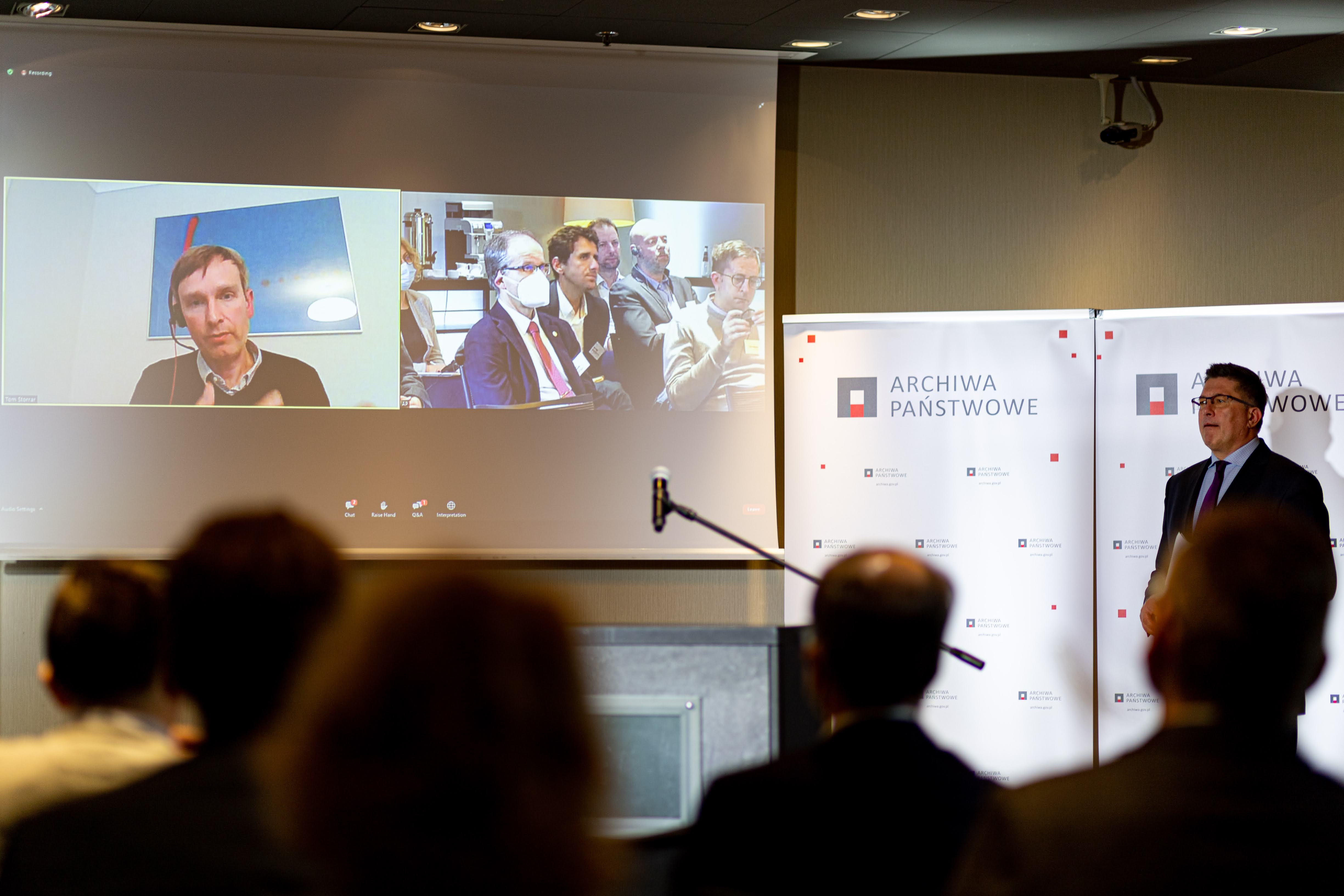 Na ekranie wystąpienie prelegenta Toma Storrara oraz z drugiej kamery obraz publiczności. Obok przy mównicy stoi prowadzący wydarzenie.