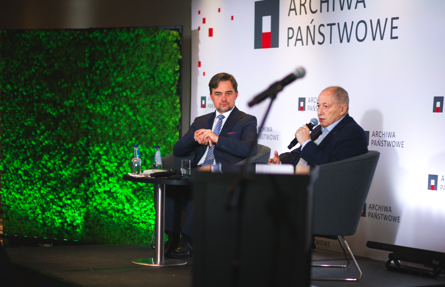 Dyrektor Pietrzyk i Józef Orzeł siedzą na fotelach. Józef Orzeł przemawia do mikrofonu. W tle ścianka z logo Archiwa Państwowe.