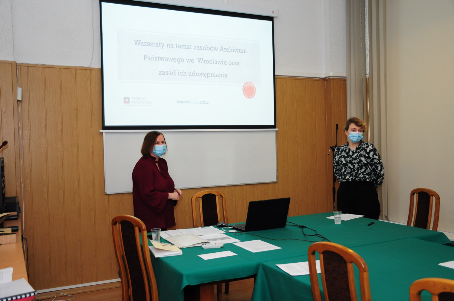 Danuta Płókarz i Karolina Rybicka stoją za stołem. Za nimi wyświetlana prezentacja pod tytułem „Warsztaty na temat zasobów Archiwum Państwowego we Wrocławiu oraz zasad ich udostępniania”.
