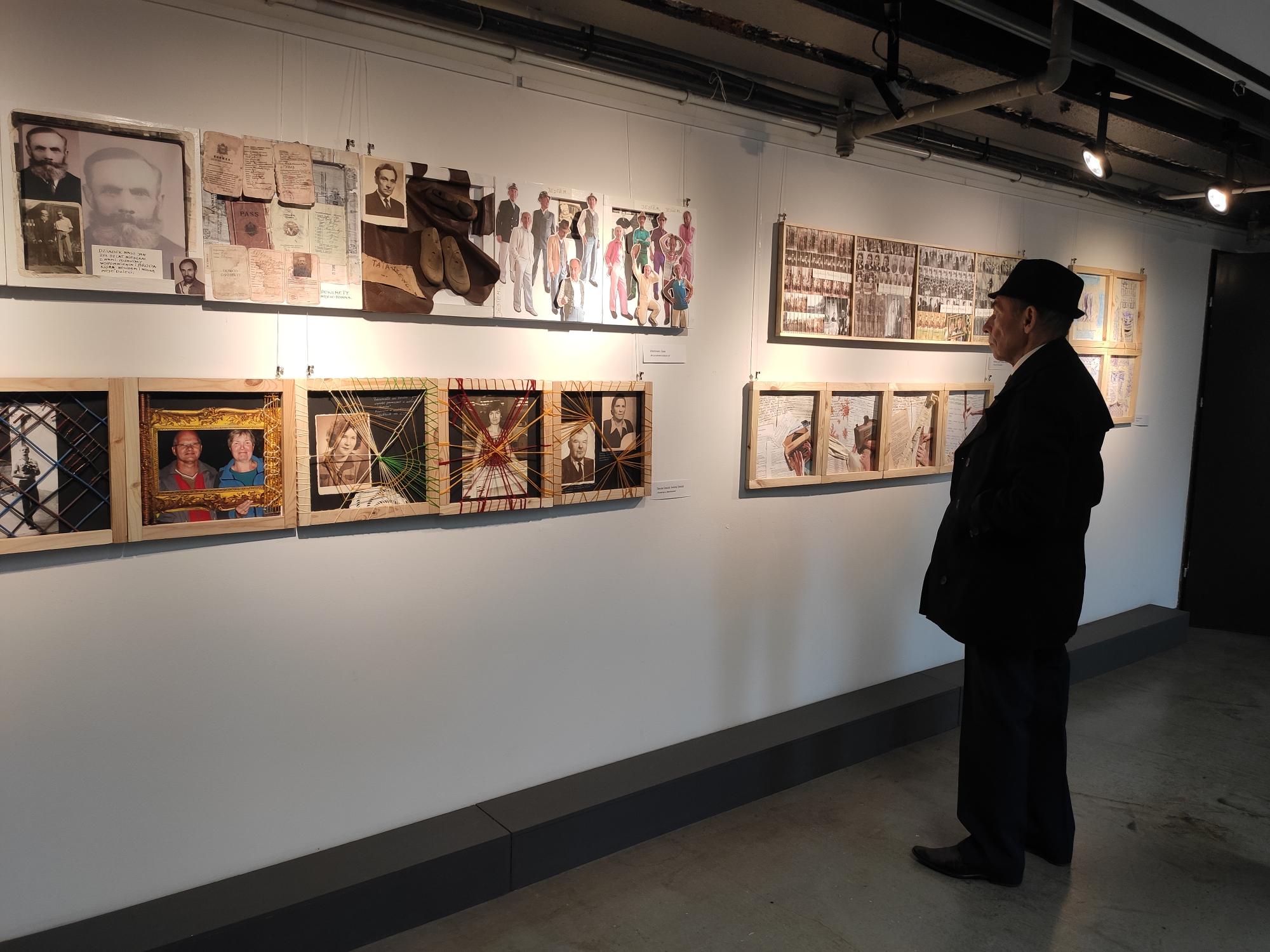 Mężczyzna w płaszczu i kapeluszu ogląda wystawę prac rozwieszonych na ścianie. 