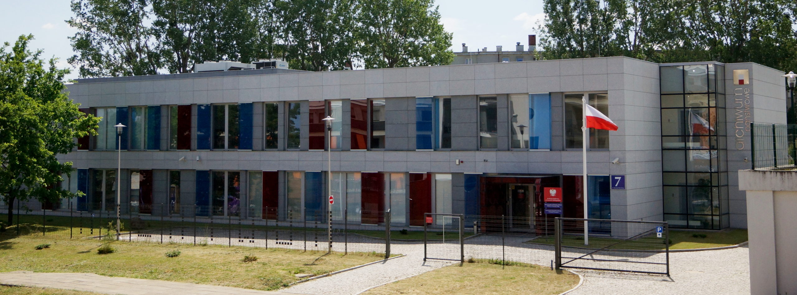 Budynek archiwum z dużą ilością przeszkleń zabarwionych na czerwono i niebiesko. Przed budynkiem trawnik oraz ogrodzenie. Na maszcie przed budynkiem powiewa polska flaga. 